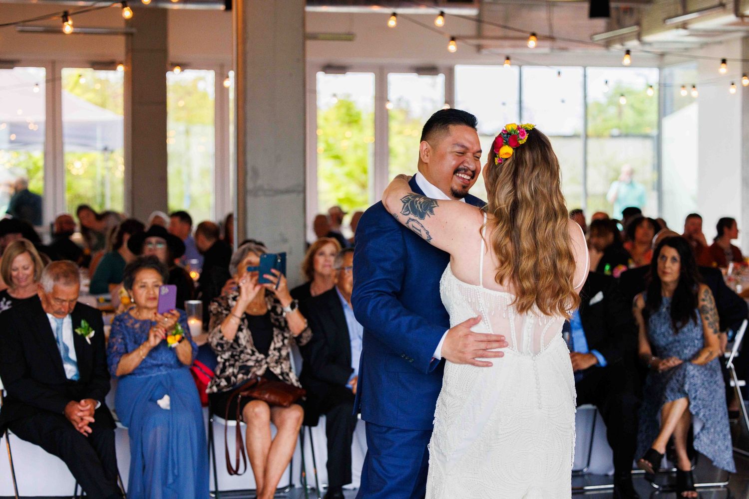 Newlyweds sharing their first dance surrounded by guests at indoor reception venue.