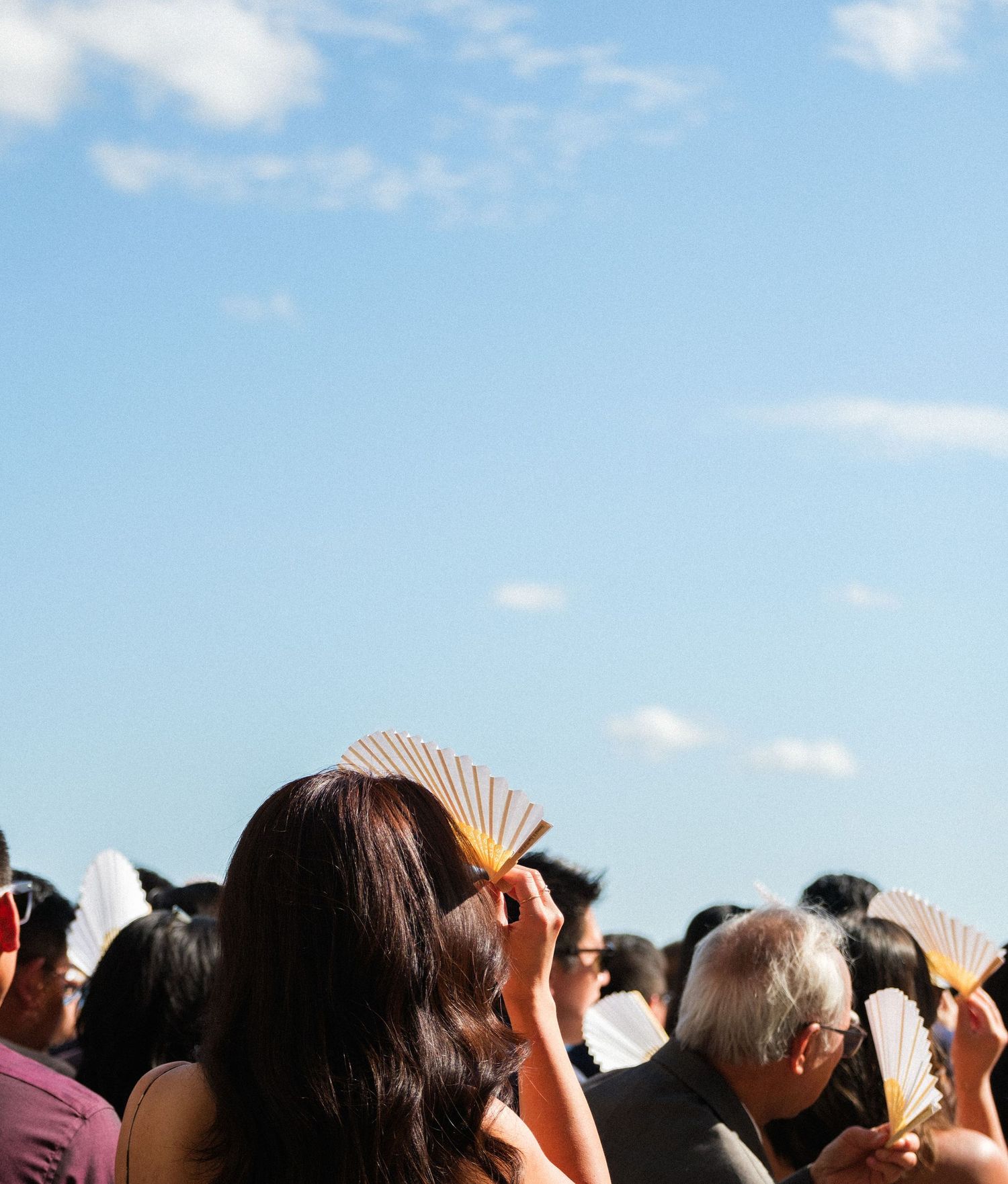Wedding guests holding paper fans against blue summer sky.