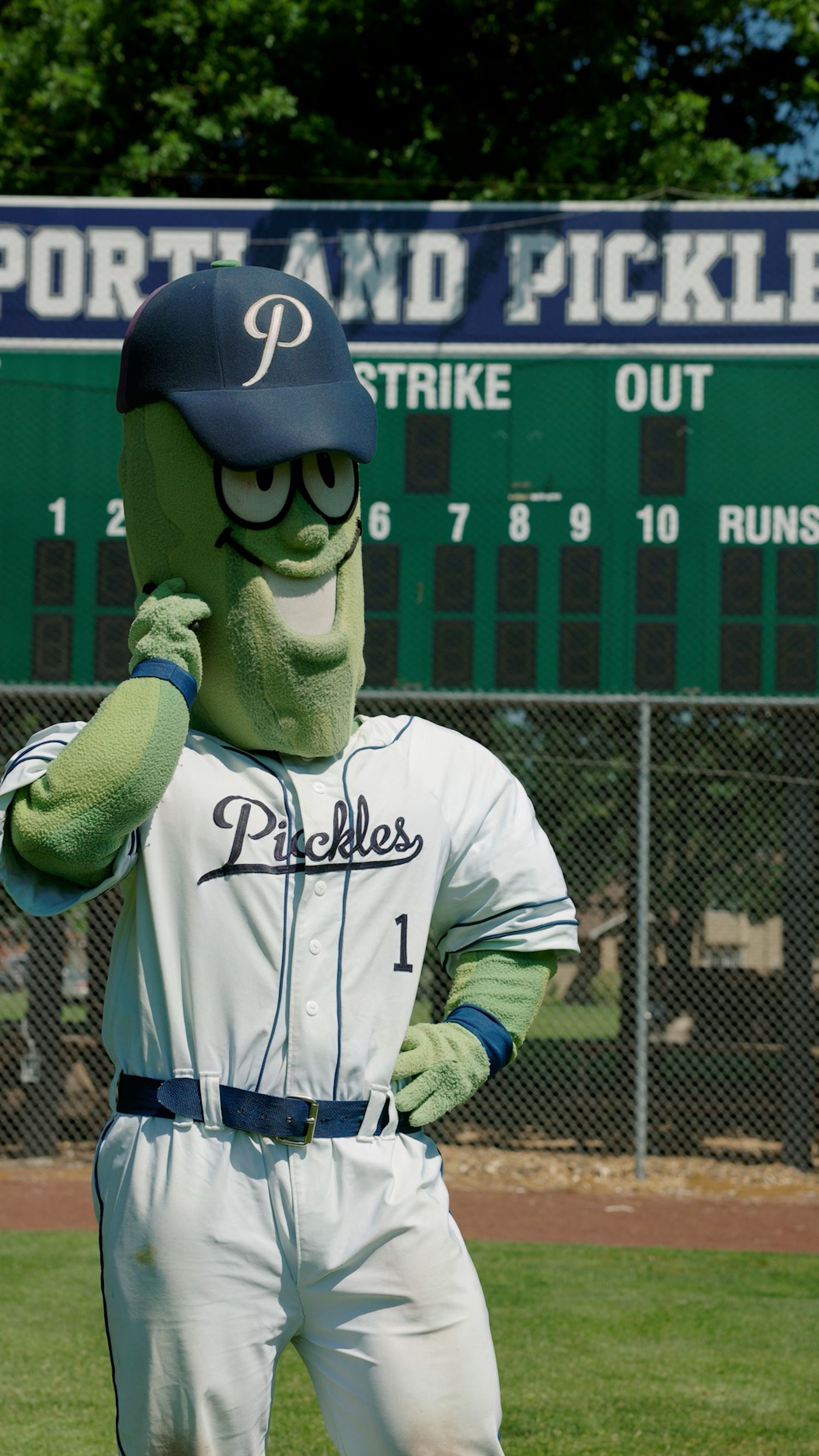 Baseball mascot Dillon the Pickle poses in Portland Pickles uniform in front of scoreboard at baseball stadium.