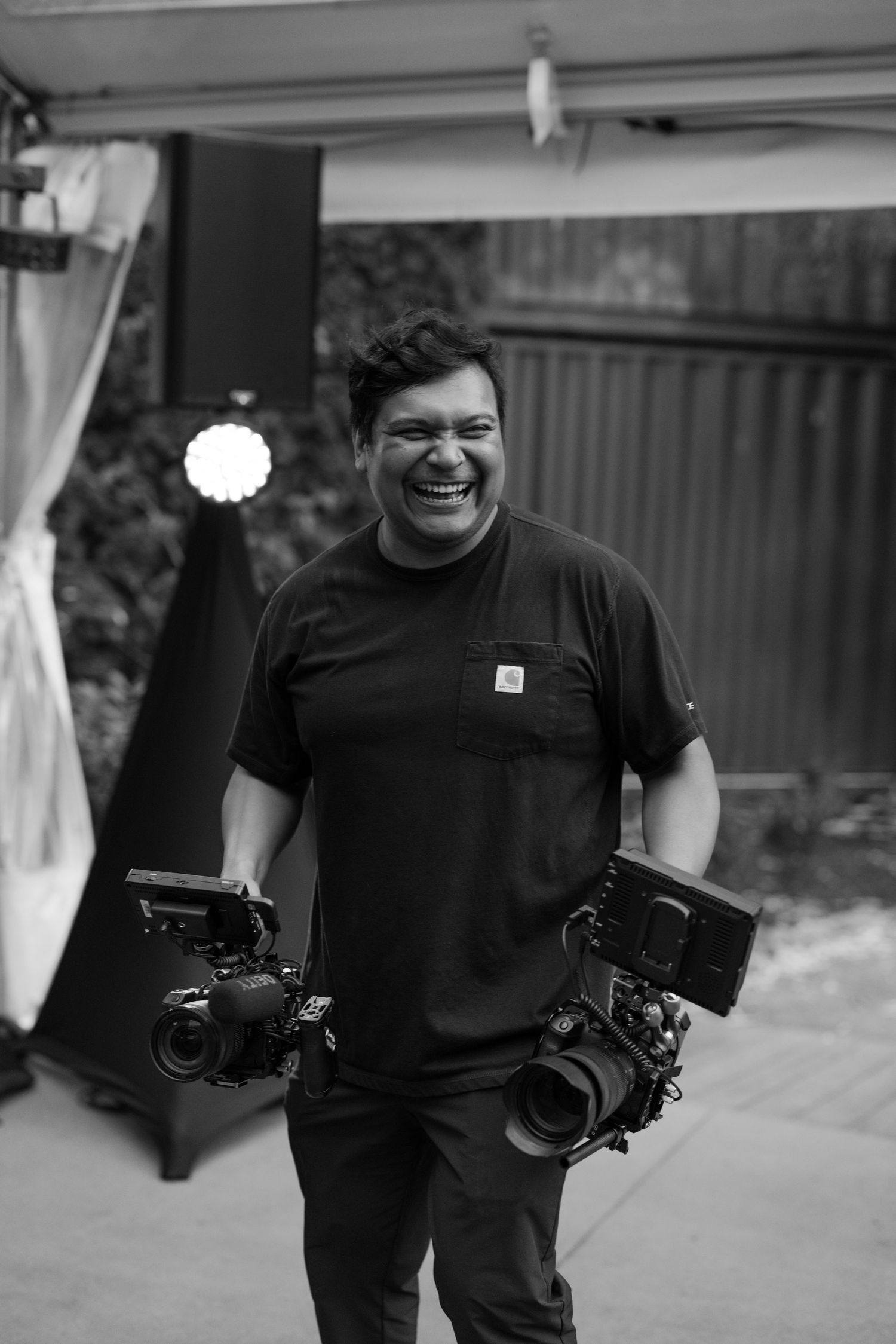 A photographer wearing a black t-shirt holds two professional cameras while smiling in a black and white photo.