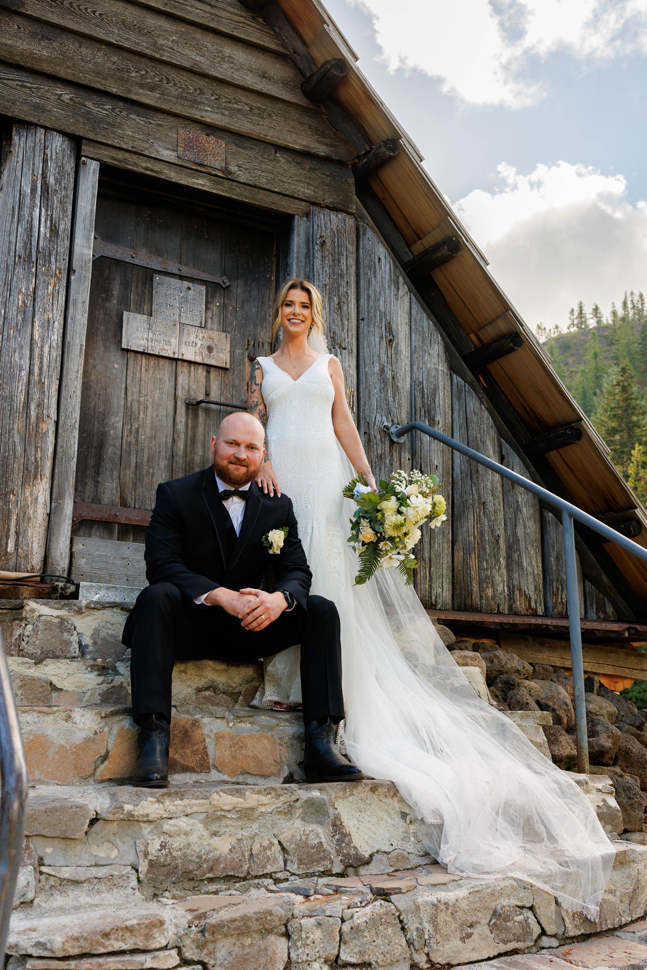 Wedding couple poses on rustic stone steps of wooden mountain cabin.