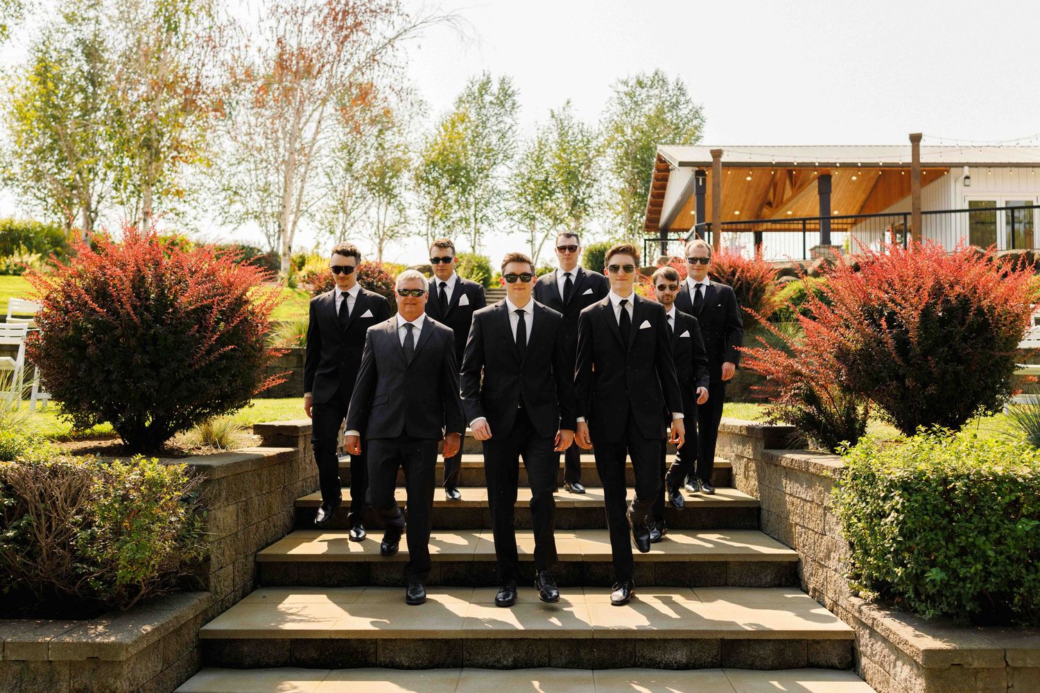 Groomsmen in black suits and ties posing on outdoor steps with autumn foliage.