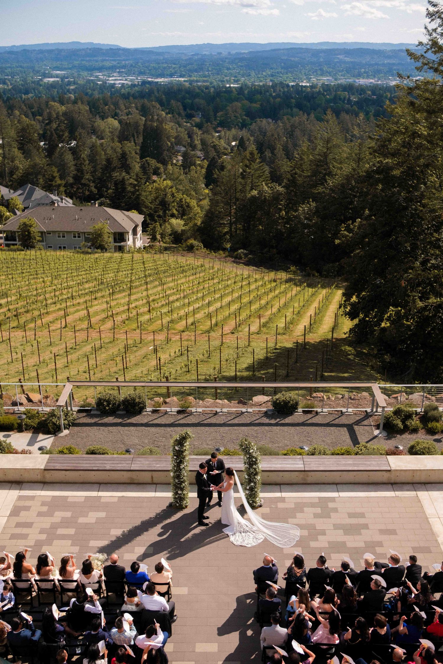 Aerial view of wedding ceremony with vineyard and forest landscape in background.
