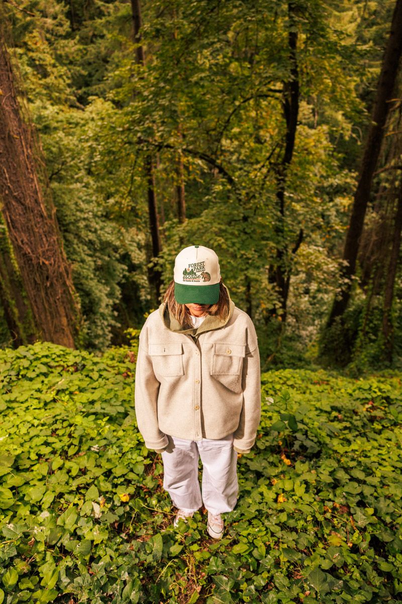 A person stands in a lush forest wearing a beige jacket, baseball cap and light colored pants surrounded by green foliage.