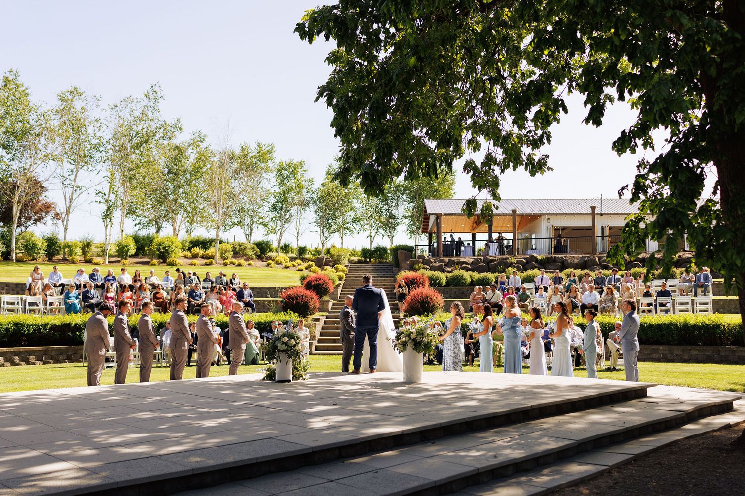 Wide angle view of outdoor wedding ceremony with guests seated under shaded trees.