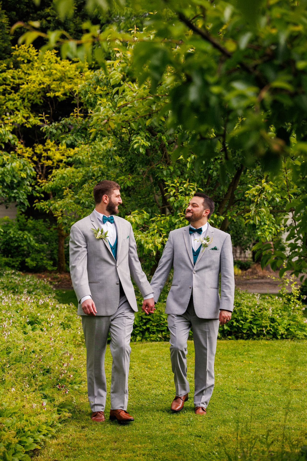 Two grooms in matching grey suits walk hand in hand through a lush garden setting.