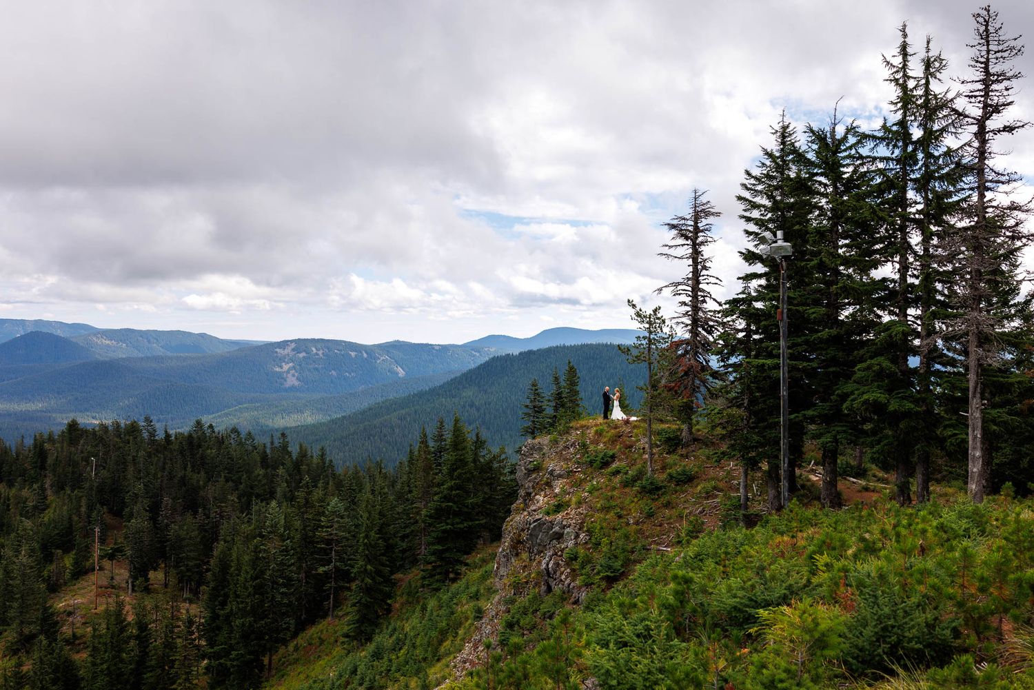 Someone stands on a scenic mountain cliff overlook surrounded by evergreen forest and mountain ranges in the distance.