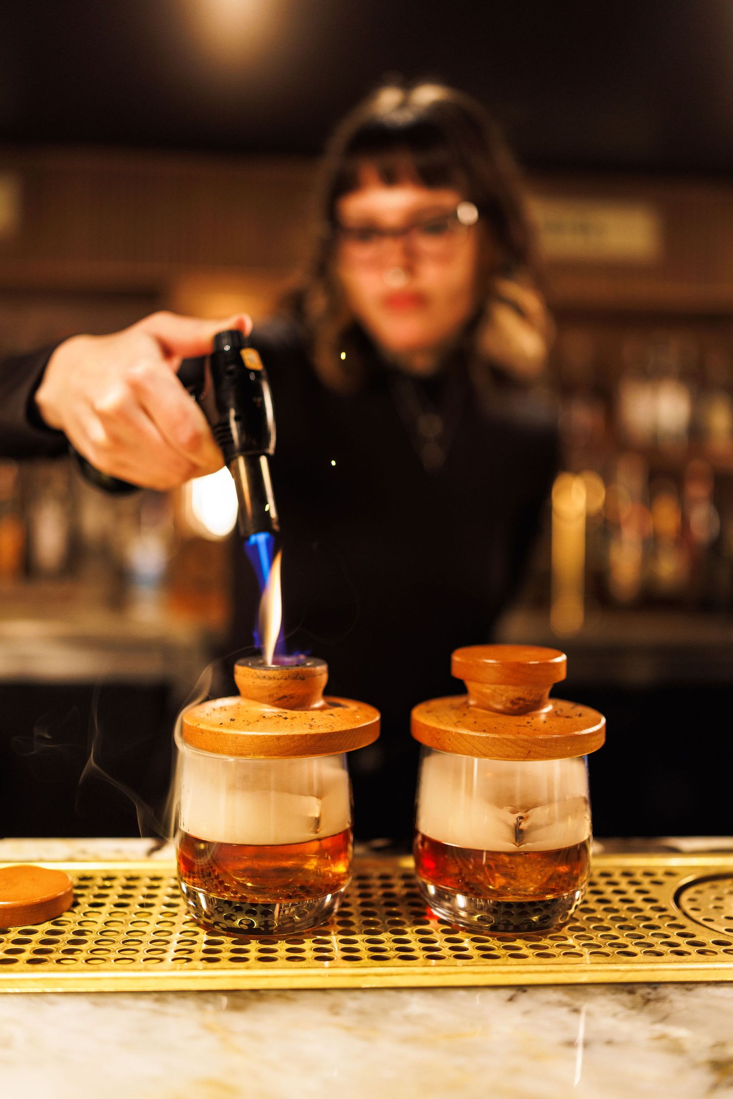 A bartender pours a smoked cocktail into two wooden-lidded glasses at an atmospheric bar counter.