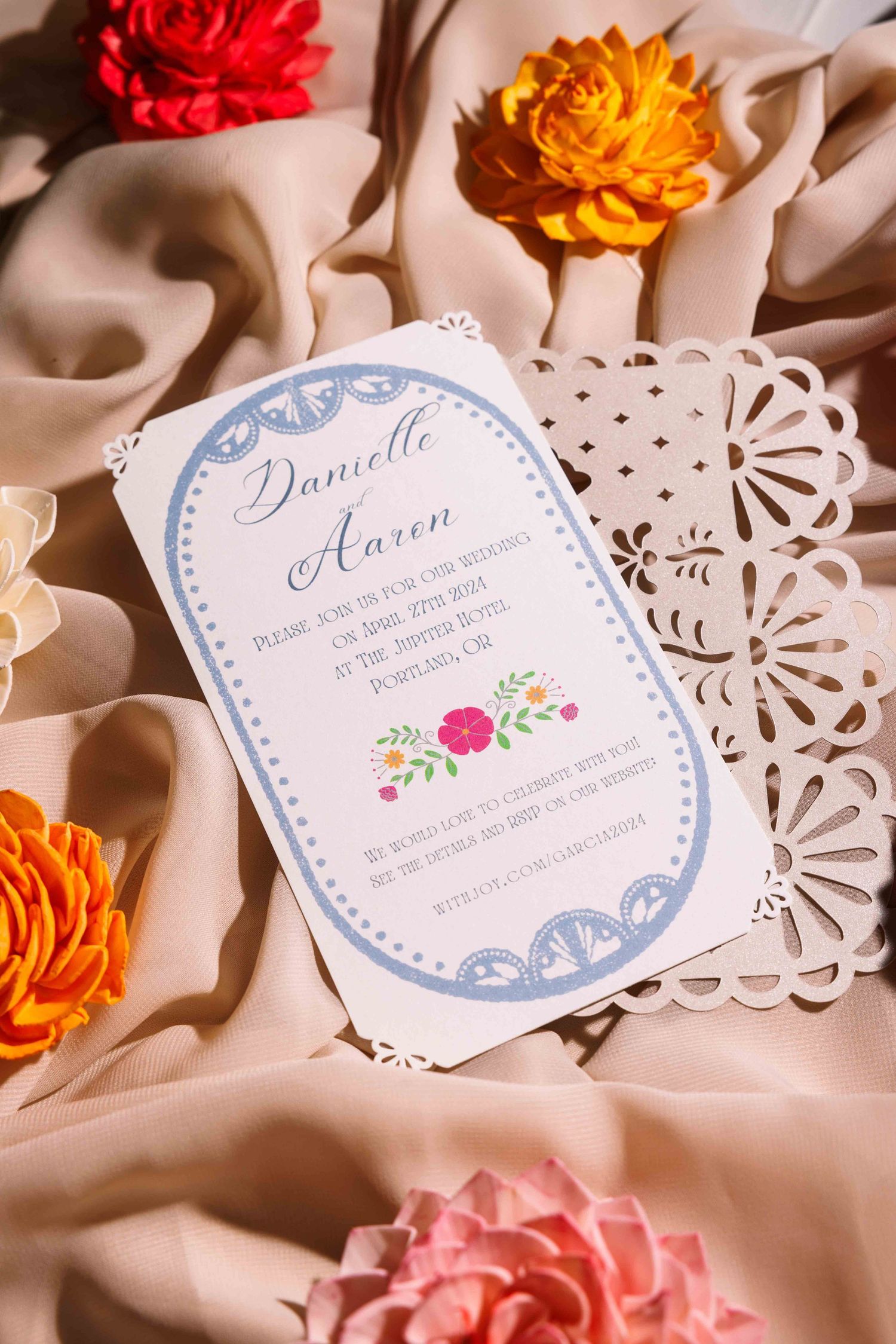 Elegant wedding invitation with lace details and floral accents surrounded by colorful paper flowers.