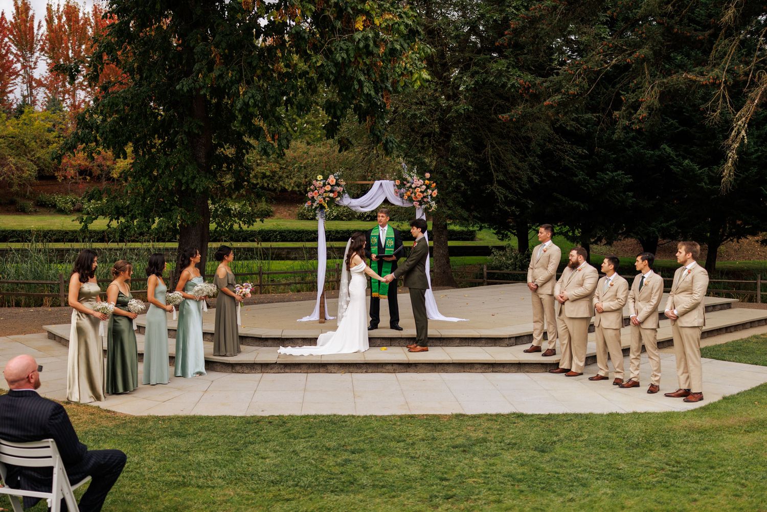Outdoor wedding ceremony with wedding party in sage green and beige attire under a decorated arch.