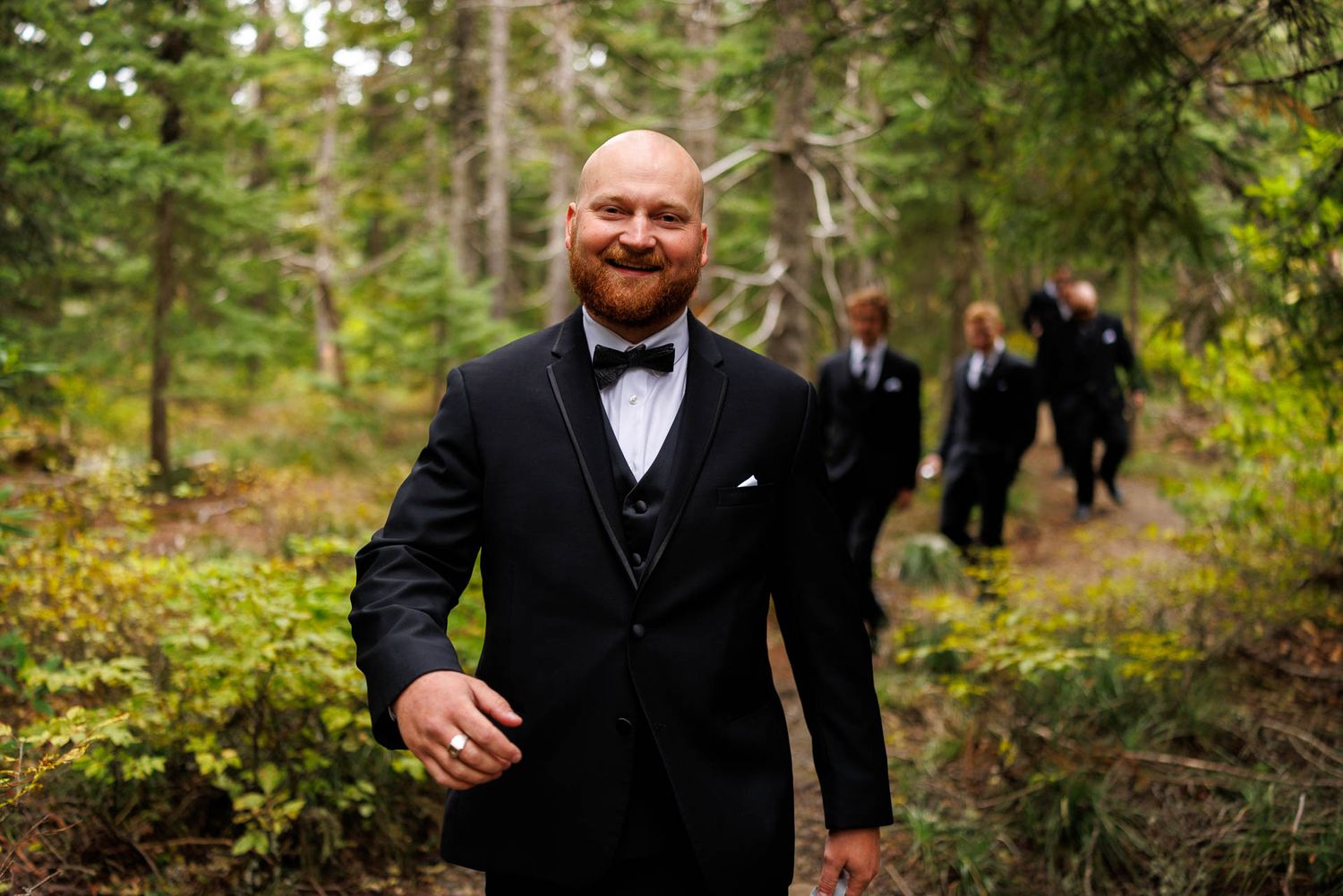 A groom in a black tuxedo smiles while walking through a forest path.