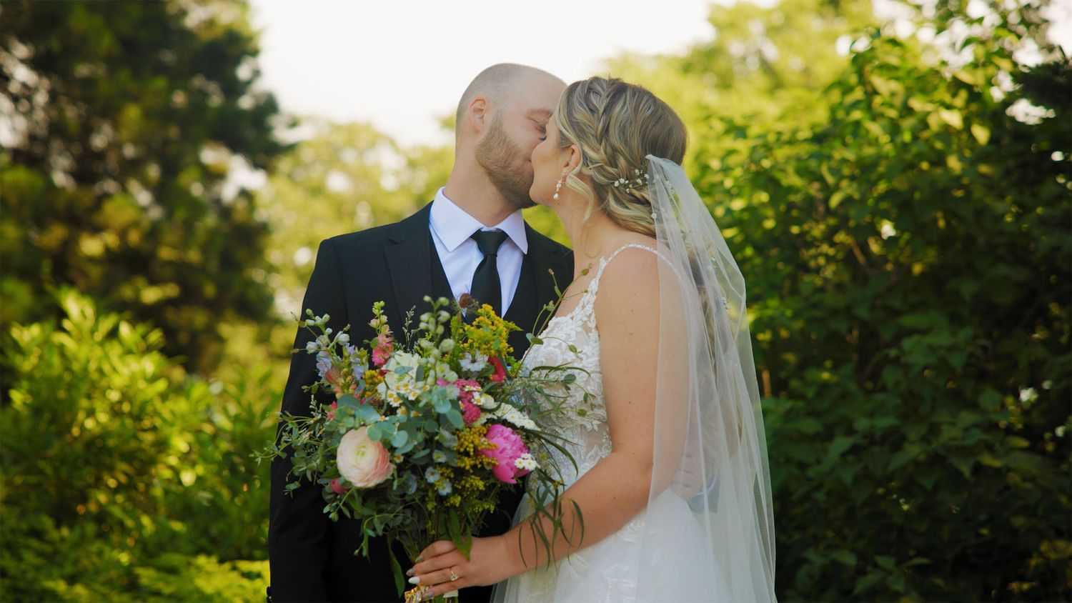 Bride and groom kissing in lush green outdoor setting at The Griffin House, holding colorful summer bouquet | Columbia River Gorge wedding
