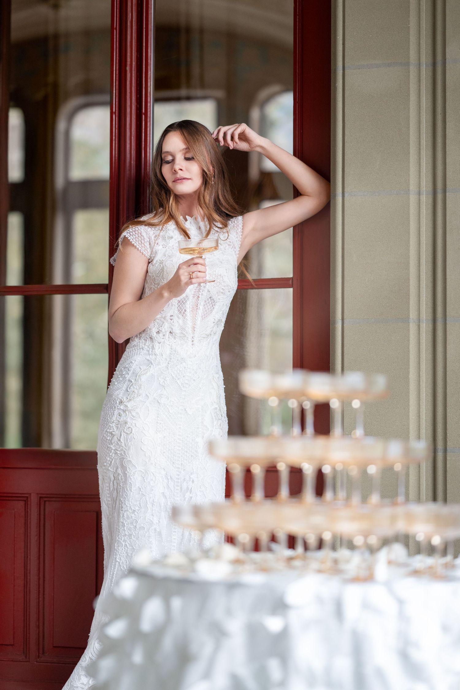 Eine Frau mit Champagnerglas in Hochzeitskleid vor einer roten Tür. Vor ihr steht eine Champagner Pyramide