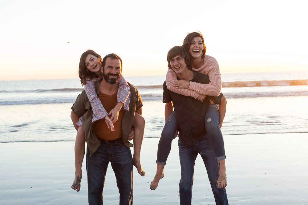 a playful family of four gives piggy back rides on the beach during a fun family portrait session on a Corona beach in San Diego, California