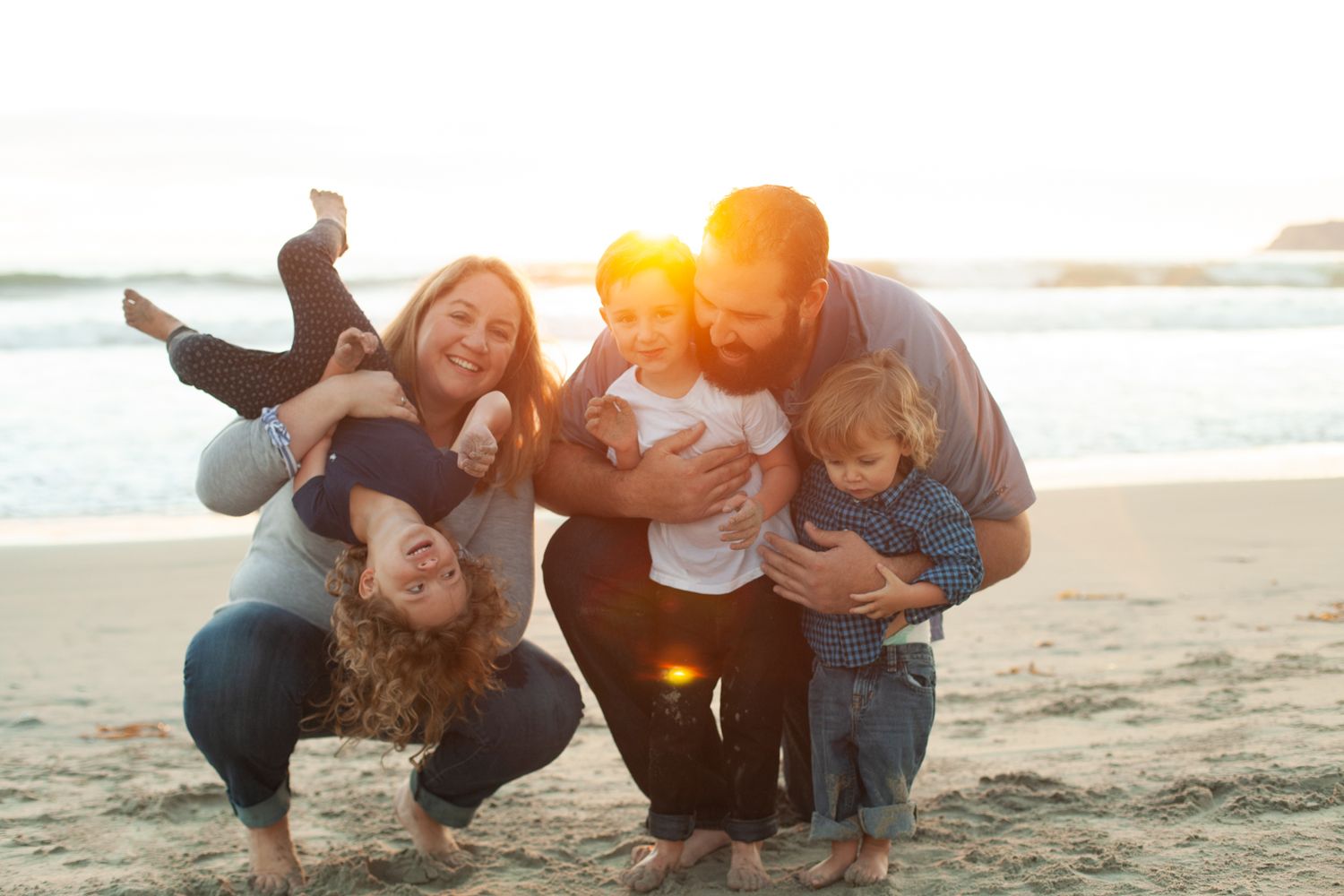 Joyful beach photo of a family of five with kids laughing and one child upside down – candid lifestyle session by the ocean