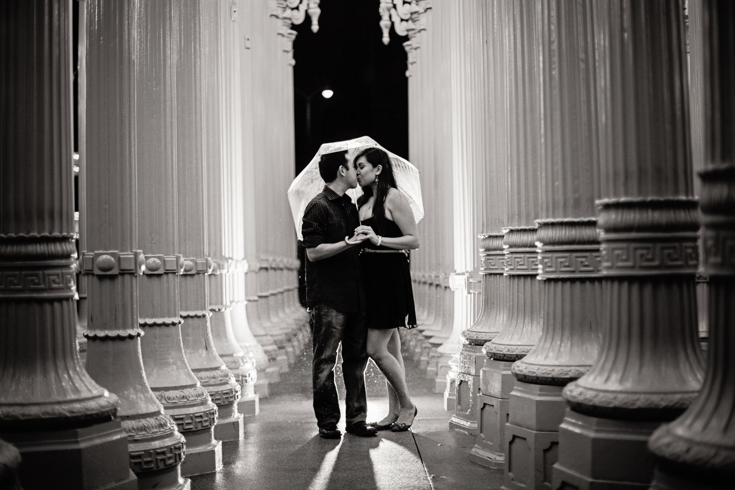 sultry black and white photo of a couple embracing under an umbrella in between the light posts at LACMA