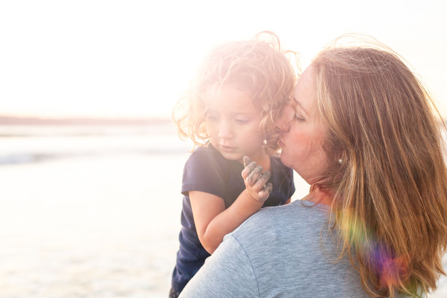 Candid photo of mom snuggling her young child on the sand at golden hour – Coronado family beach session