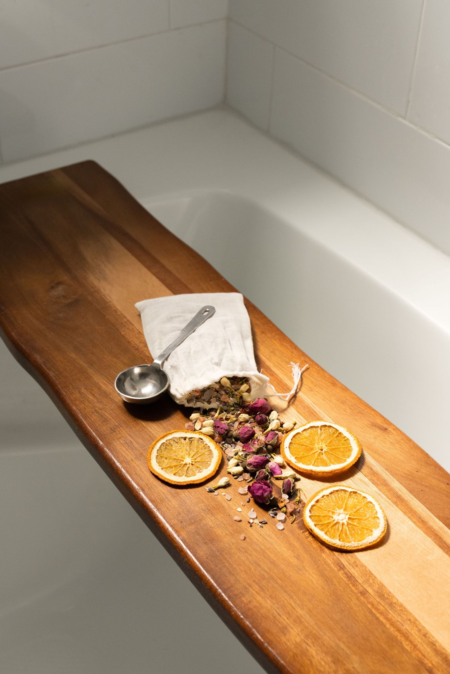 Dried orange slices and flower petals scattered on wooden bath caddy with silver spoon and white cloth.