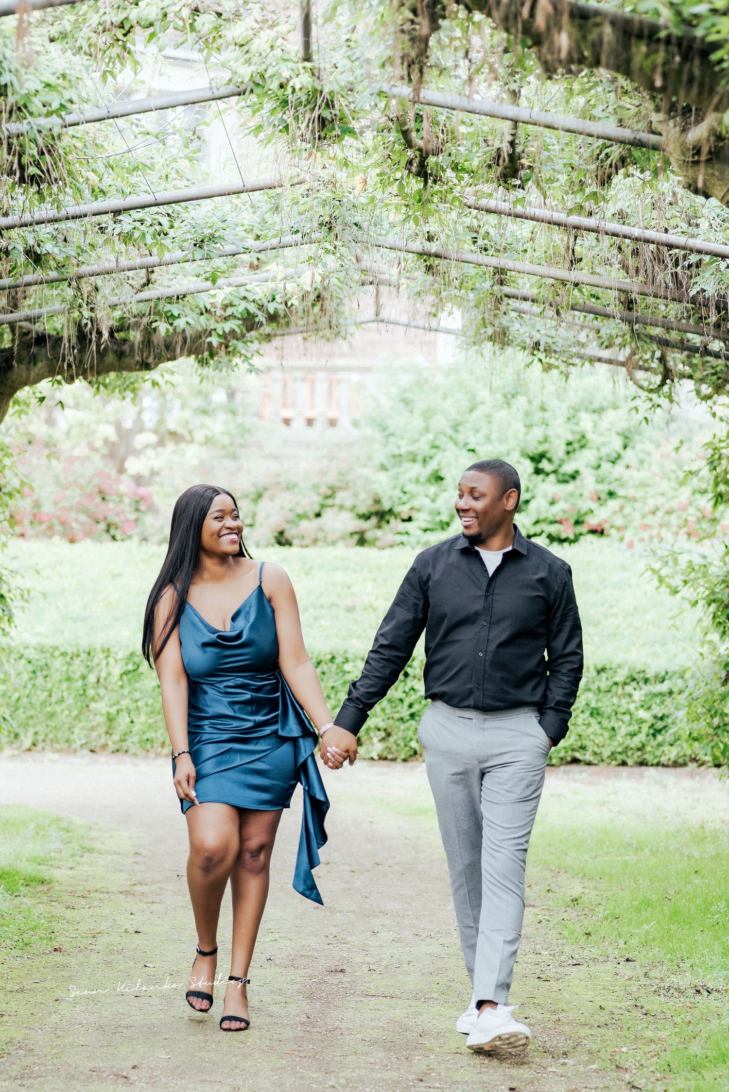 A couple holding hands while walking beneath a vine-covered garden archway in a lush outdoor setting.