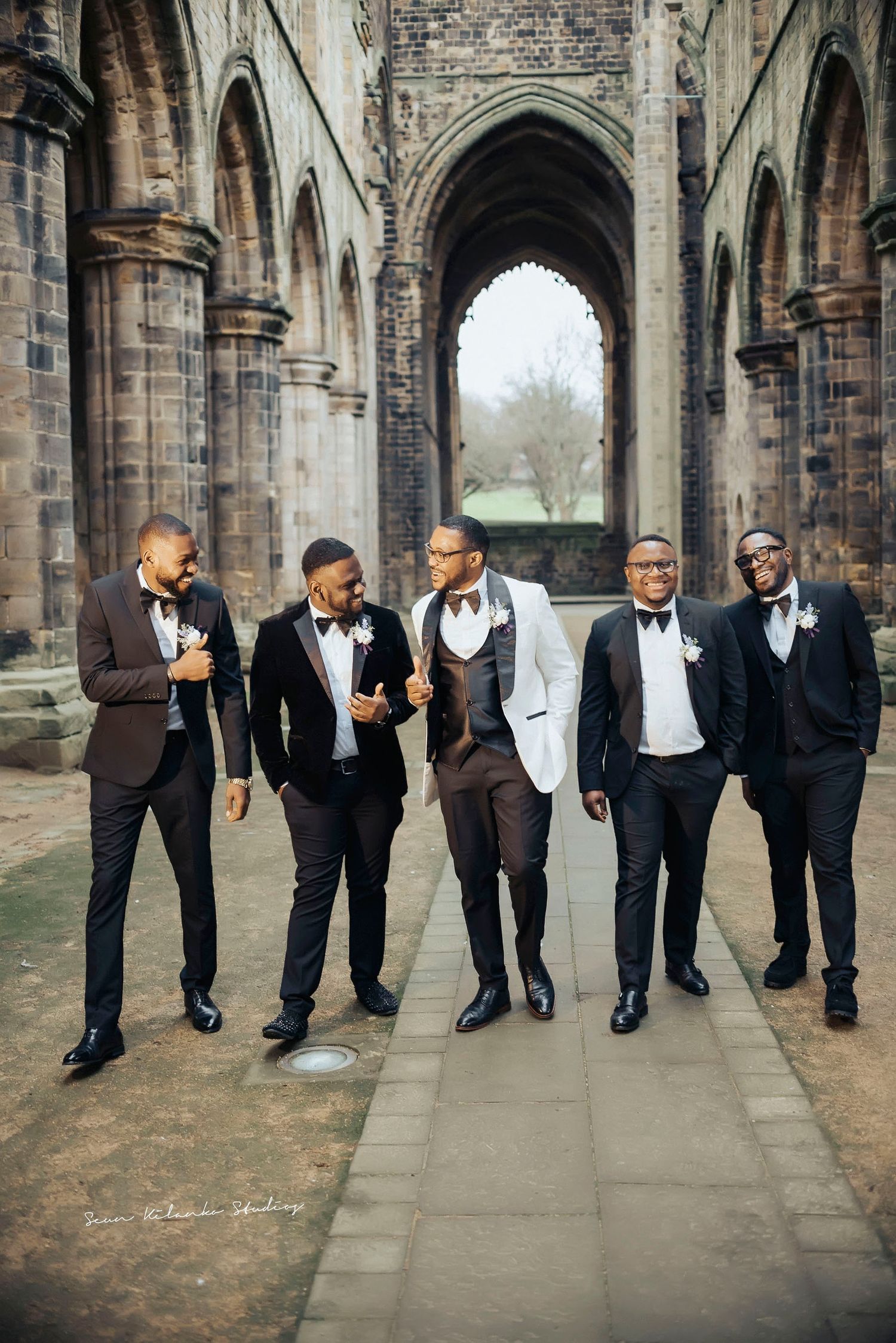 A group of groomsmen in formal attire walking through a historic stone archway.
