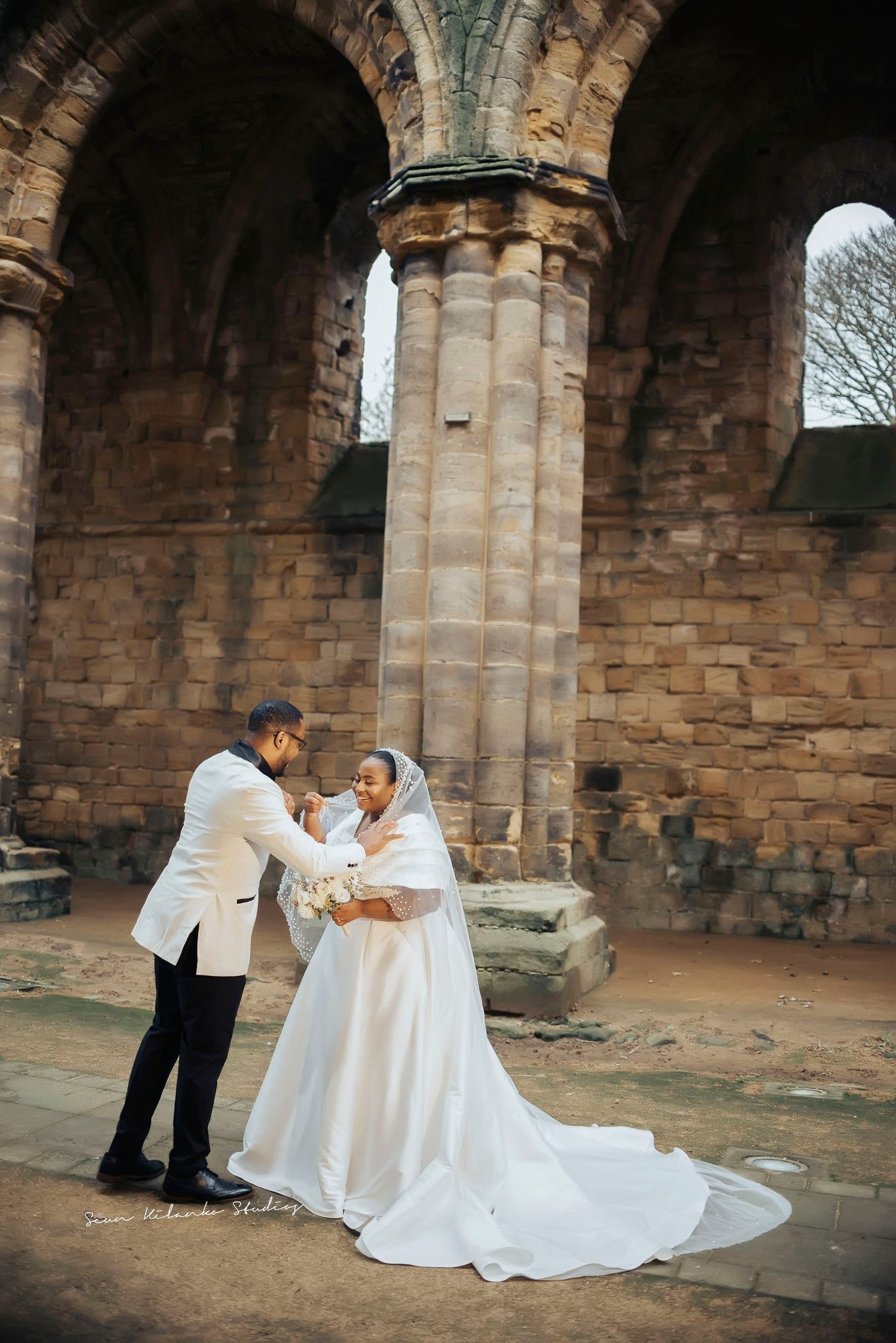 A wedding couple shares a romantic moment under historic stone arches.