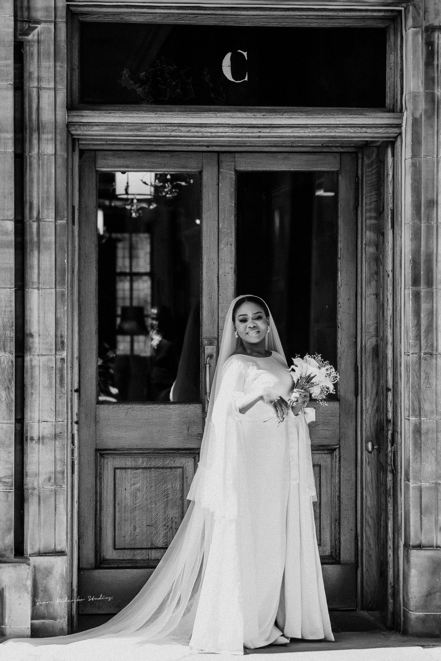 A bride in an elegant white gown poses in a classic black and white portrait at a doorway.