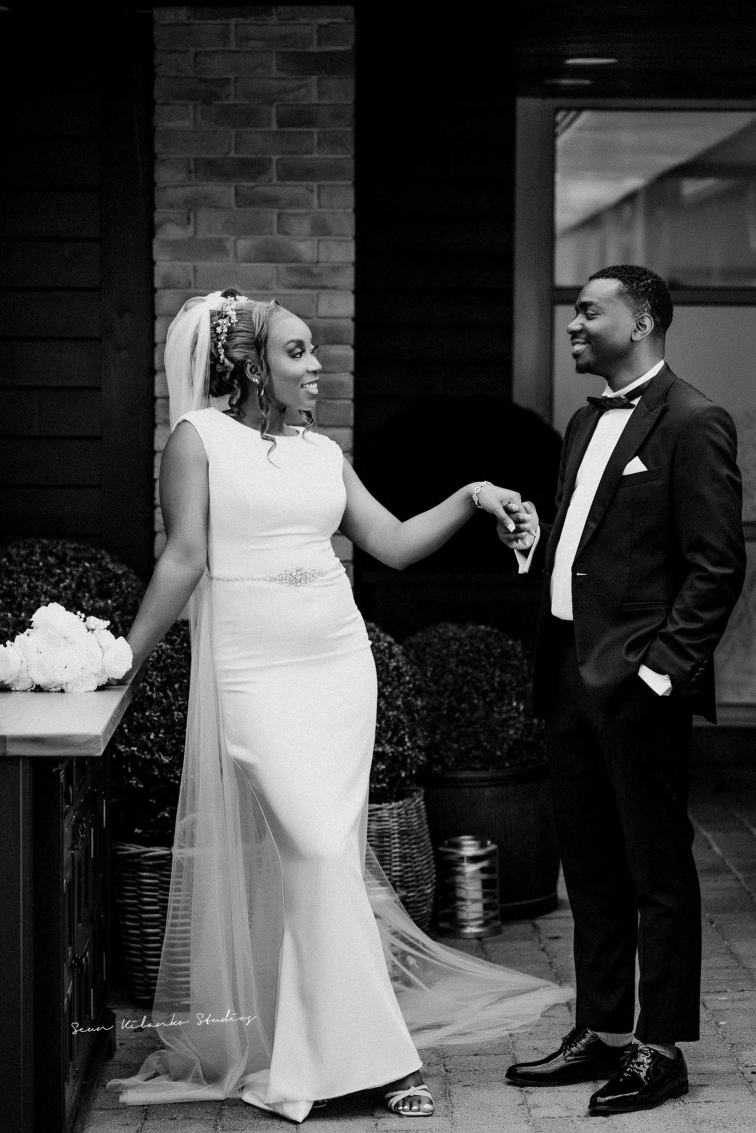 Elegant wedding couple poses in formal attire against a brick wall backdrop.