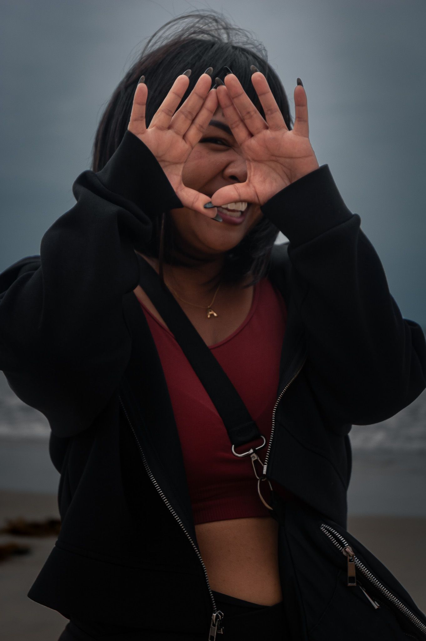 Person in black hoodie and red crop top playfully covering face with hands while smiling against moody background.