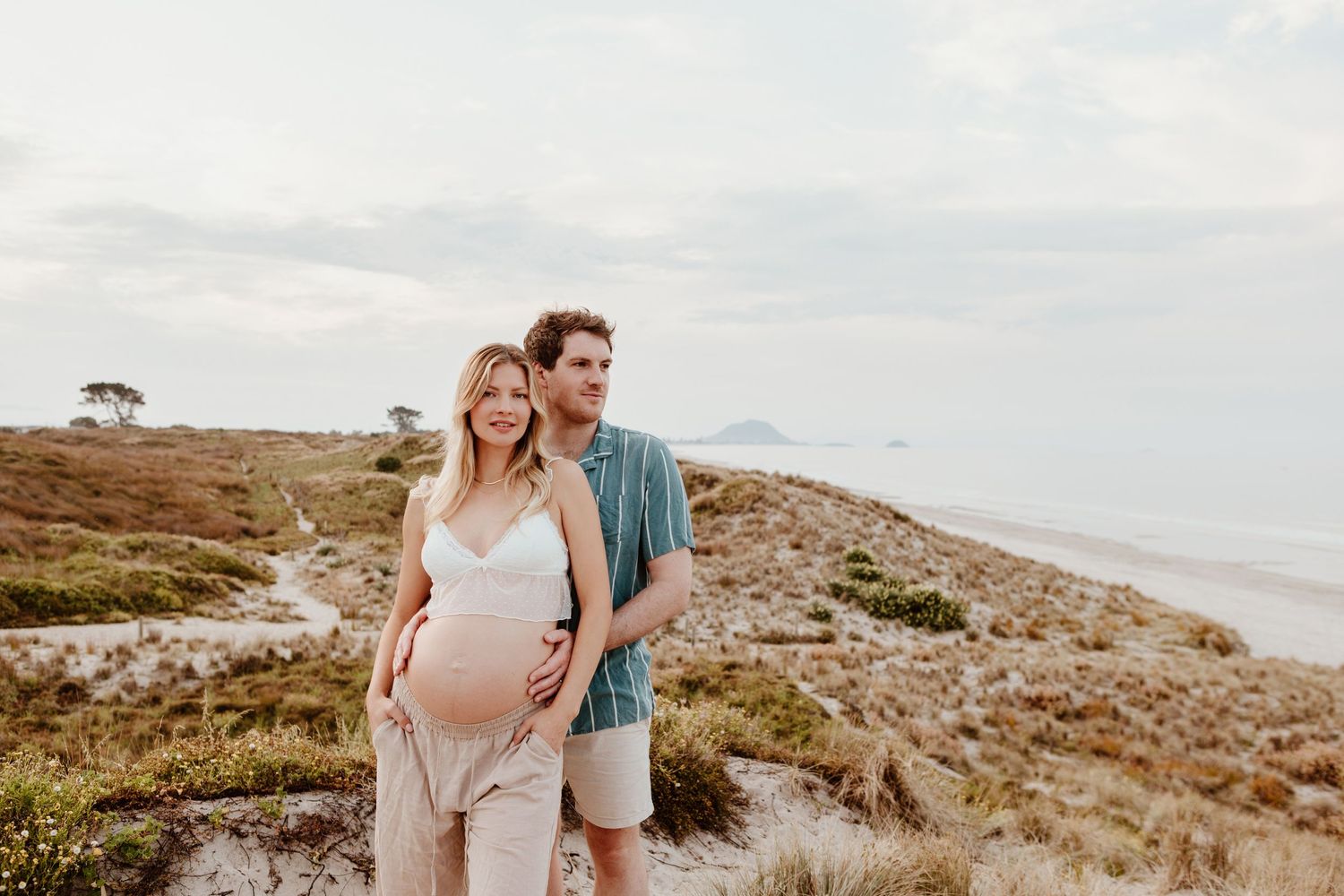 A couple stands on a coastal hillside at sunset with ocean views in the background and dunes surrounding them. Papamoa photographer