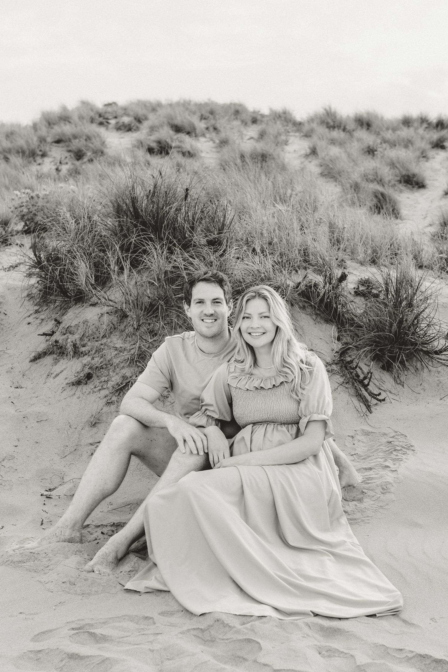 Couple sitting together on sandy beach dunes in black and white photograph.