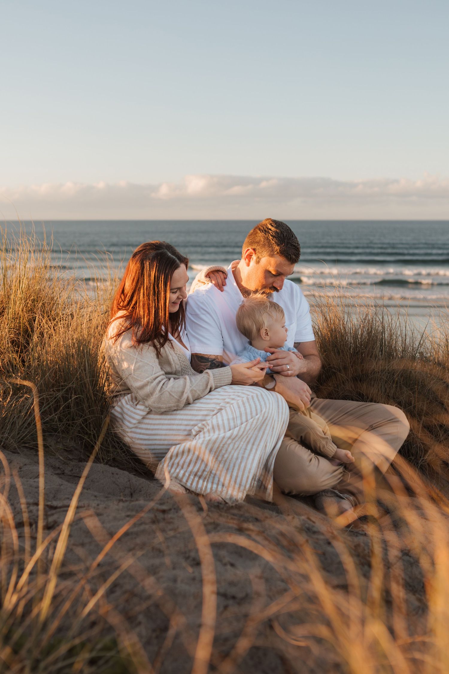 Family shares tender moment together while sitting in beach grass during golden hour sunset.