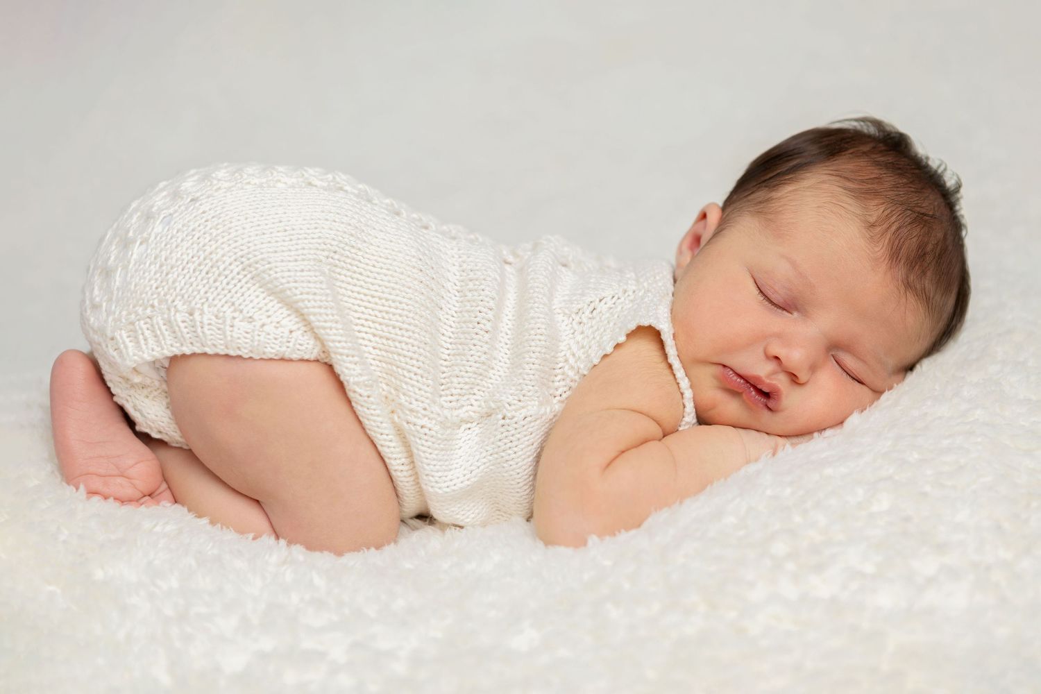Baby in white knit outfit sleeping soundly on textured white bedding. Baby photographer Tauranga