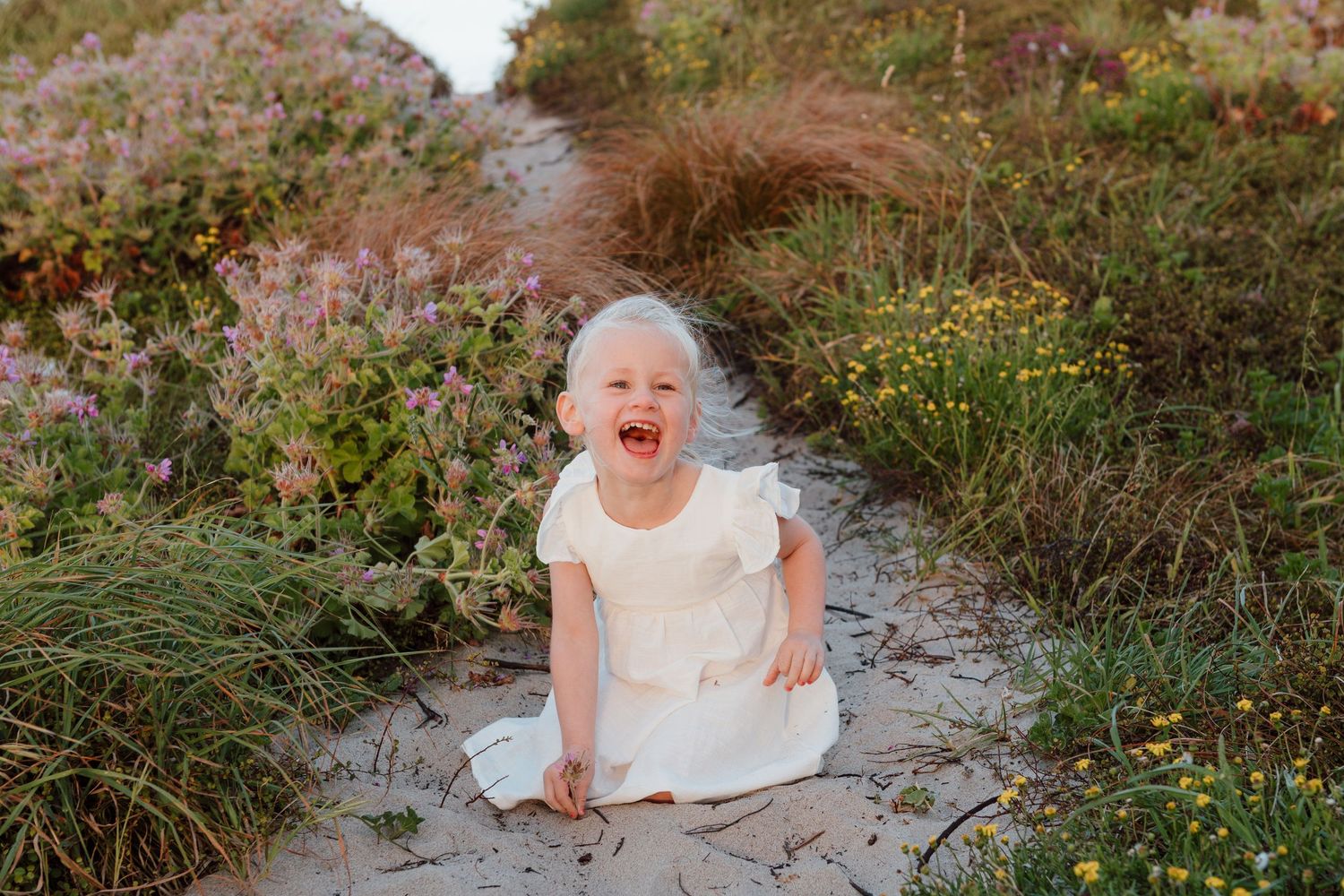 A laughing child in a white dress sitting on a sandy beach path.