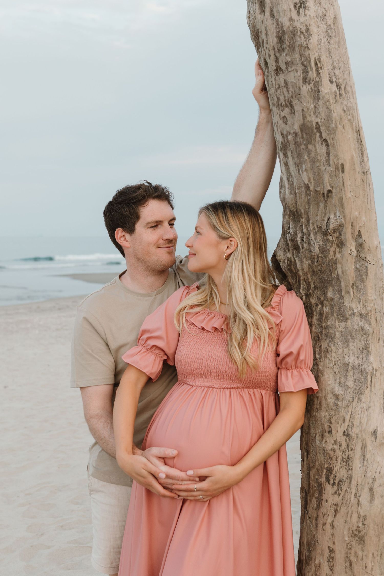 Expectant couple embracing by driftwood tree on beach at sunset.