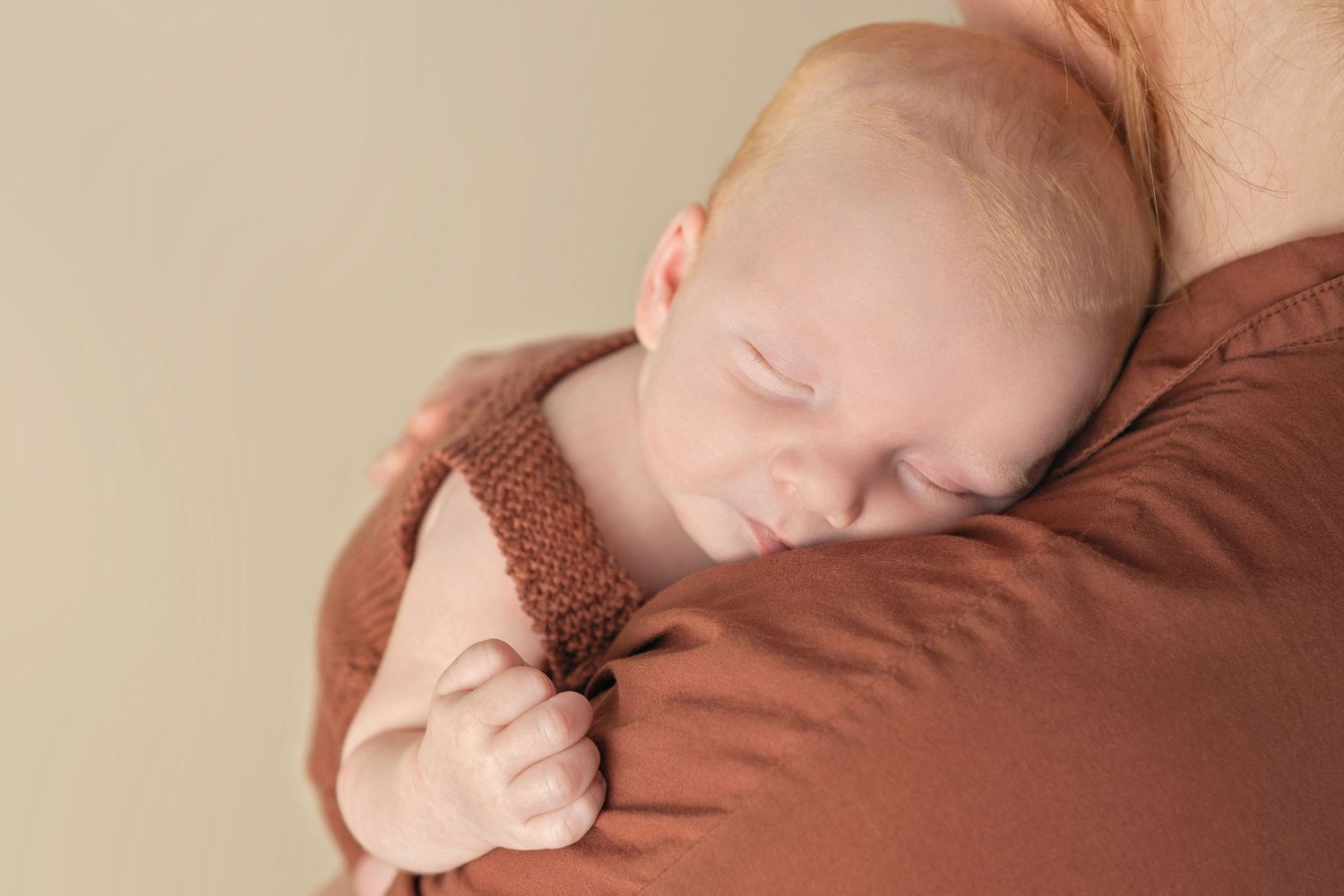 A peaceful sleeping newborn in brown knit clothing resting against someone.