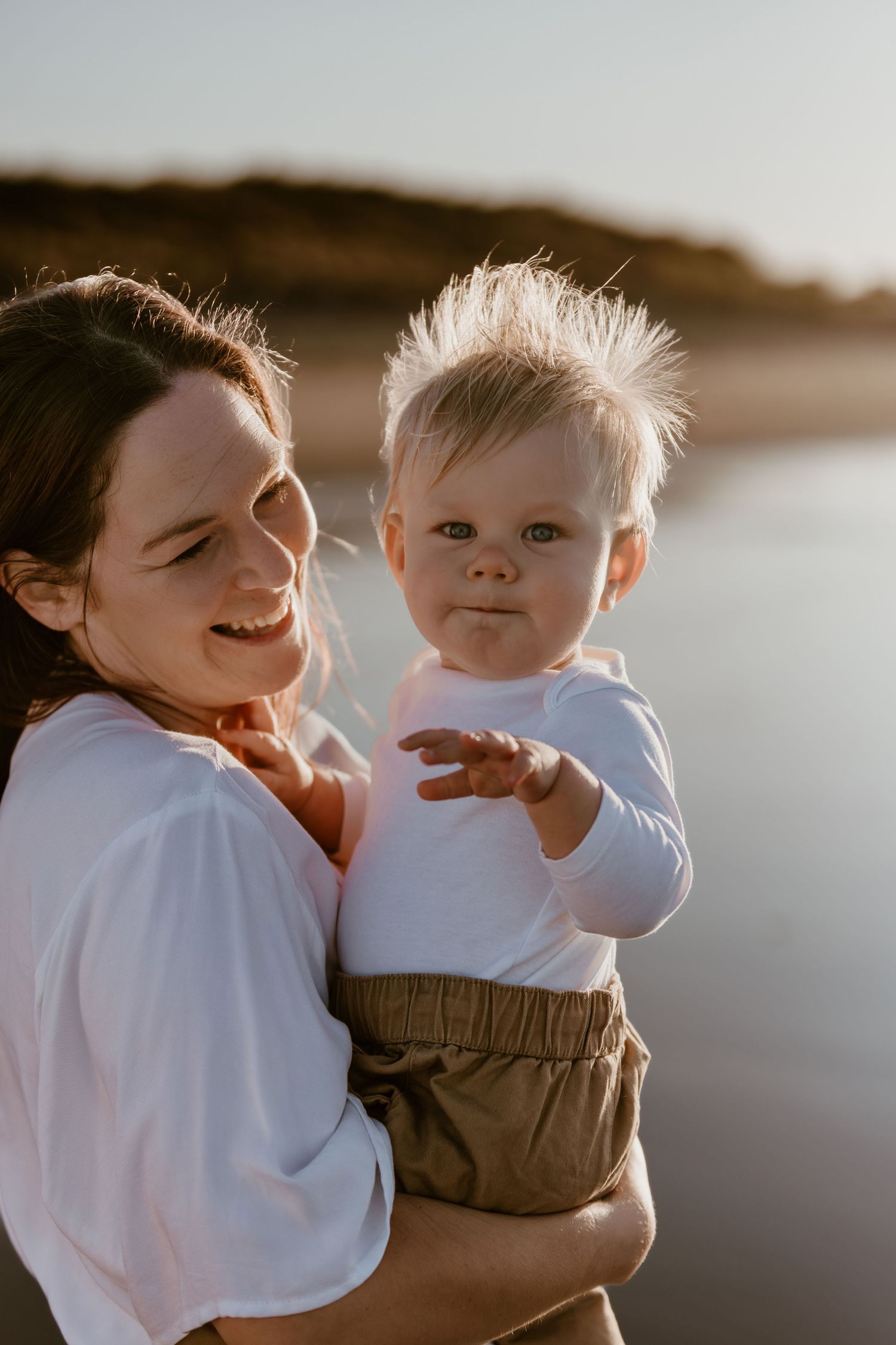 Mom and baby share joyful moment together against sunset beach backdrop.