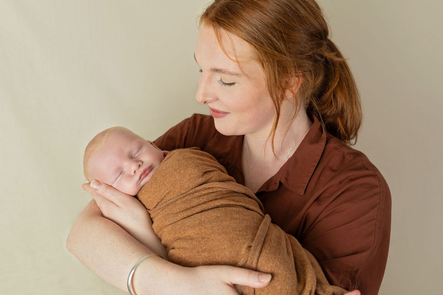A tender moment between parent and infant wrapped in a brown swaddle against neutral backdrop.