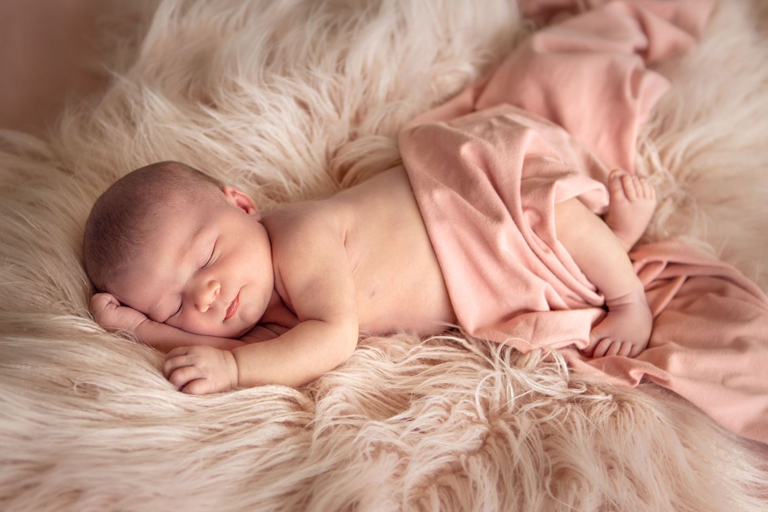 Sleeping newborn on soft pink fur blanket in studio lighting.