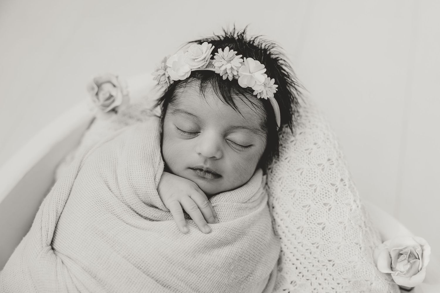 A sleeping newborn wearing a white floral crown wrapped in a delicate white blanket.