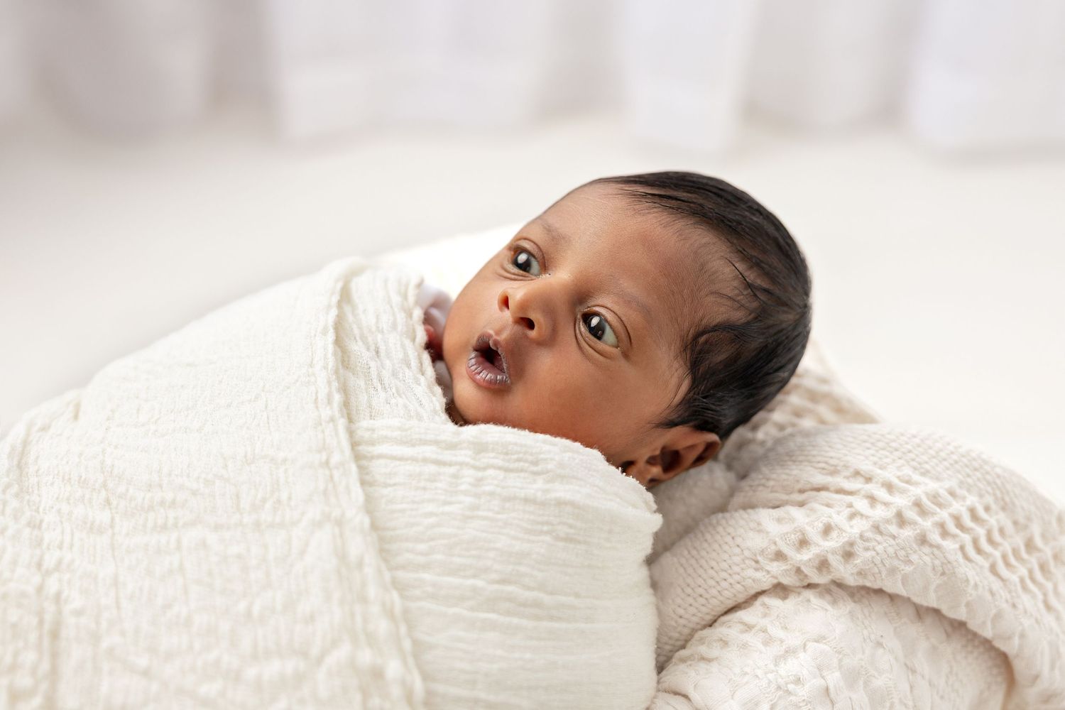 Baby swaddled in white blanket looking at camera with curious expression.