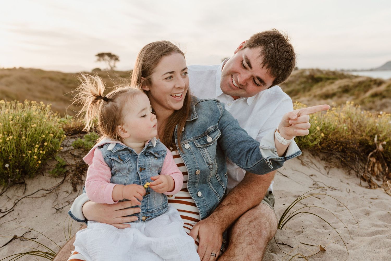 Family enjoys a sunset moment together sitting on sandy beach dunes surrounded by coastal grasses and wildflowers. Papamoa beach photos