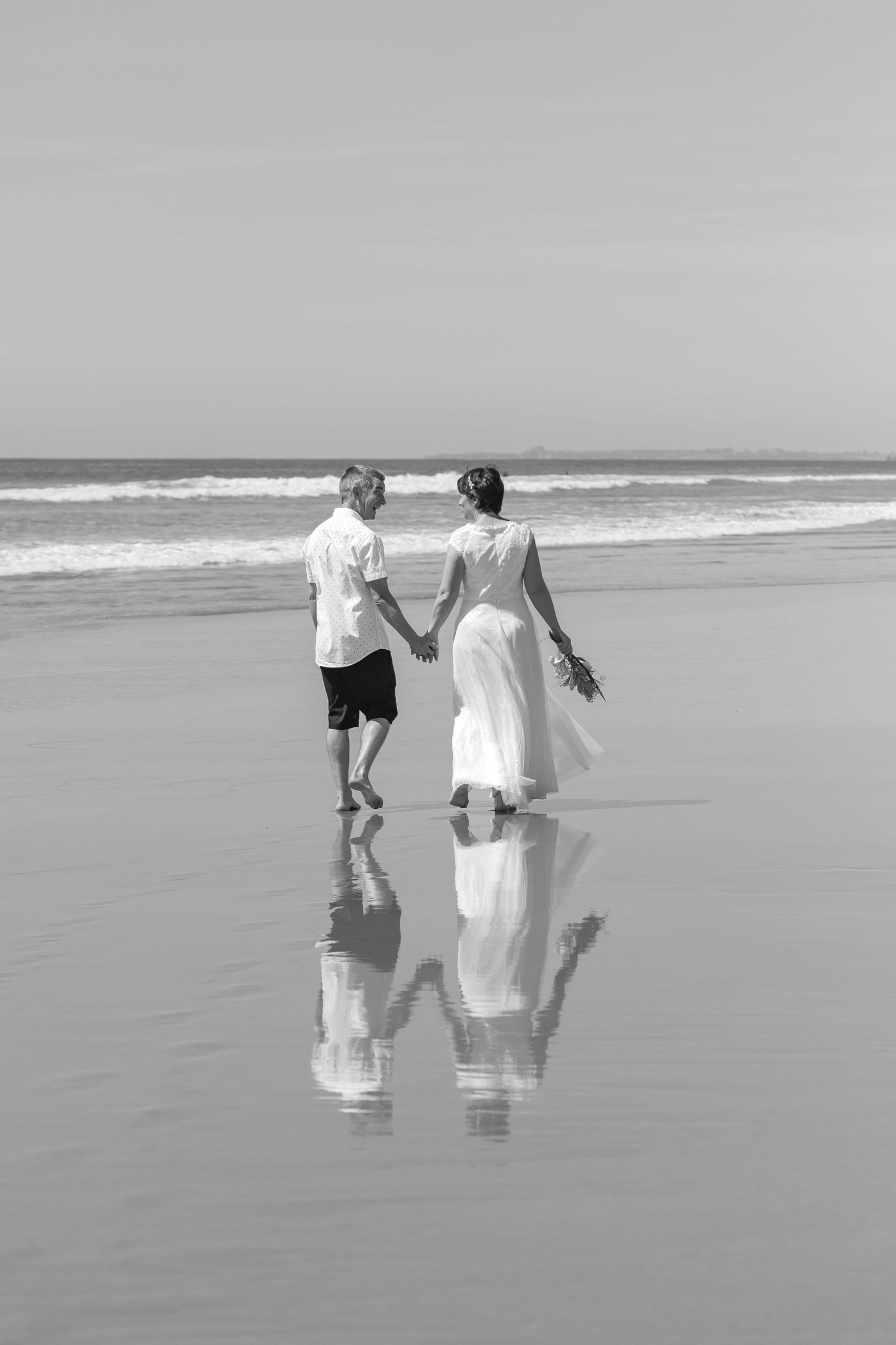 A couple walking hand in hand on a reflective beach during an overcast day, shown in black and white.