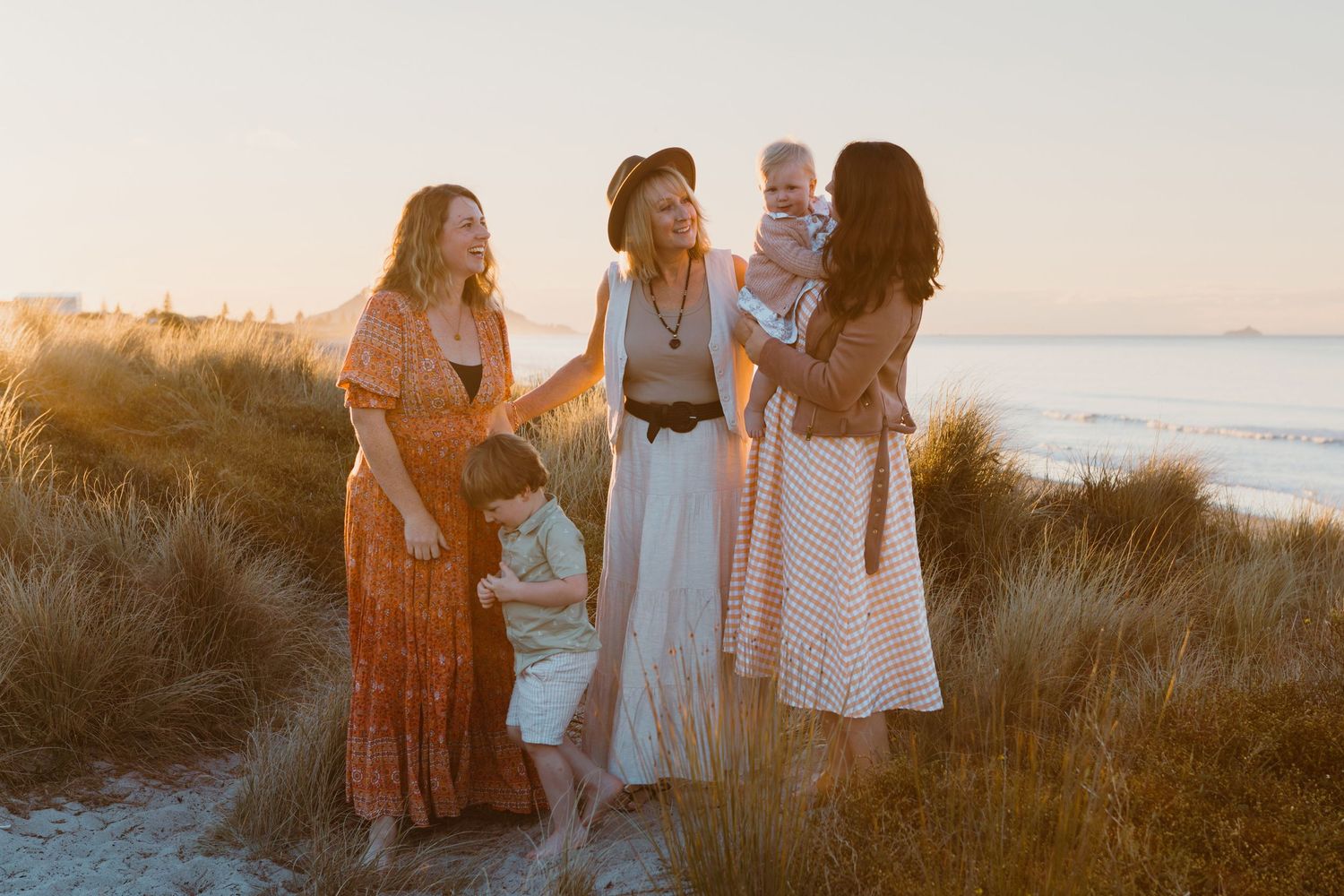 Three women in flowing dresses share a sunset moment together on coastal dunes overlooking the ocean.