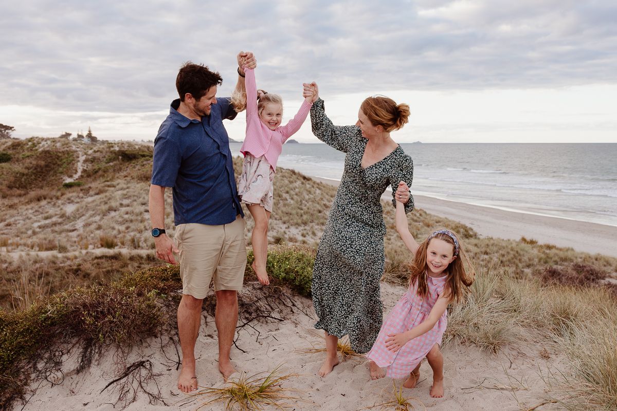 Joyful family plays and dances together on coastal sand dunes at sunset by the ocean. Mount Maunganui sunset photoshoot. Local Photographer