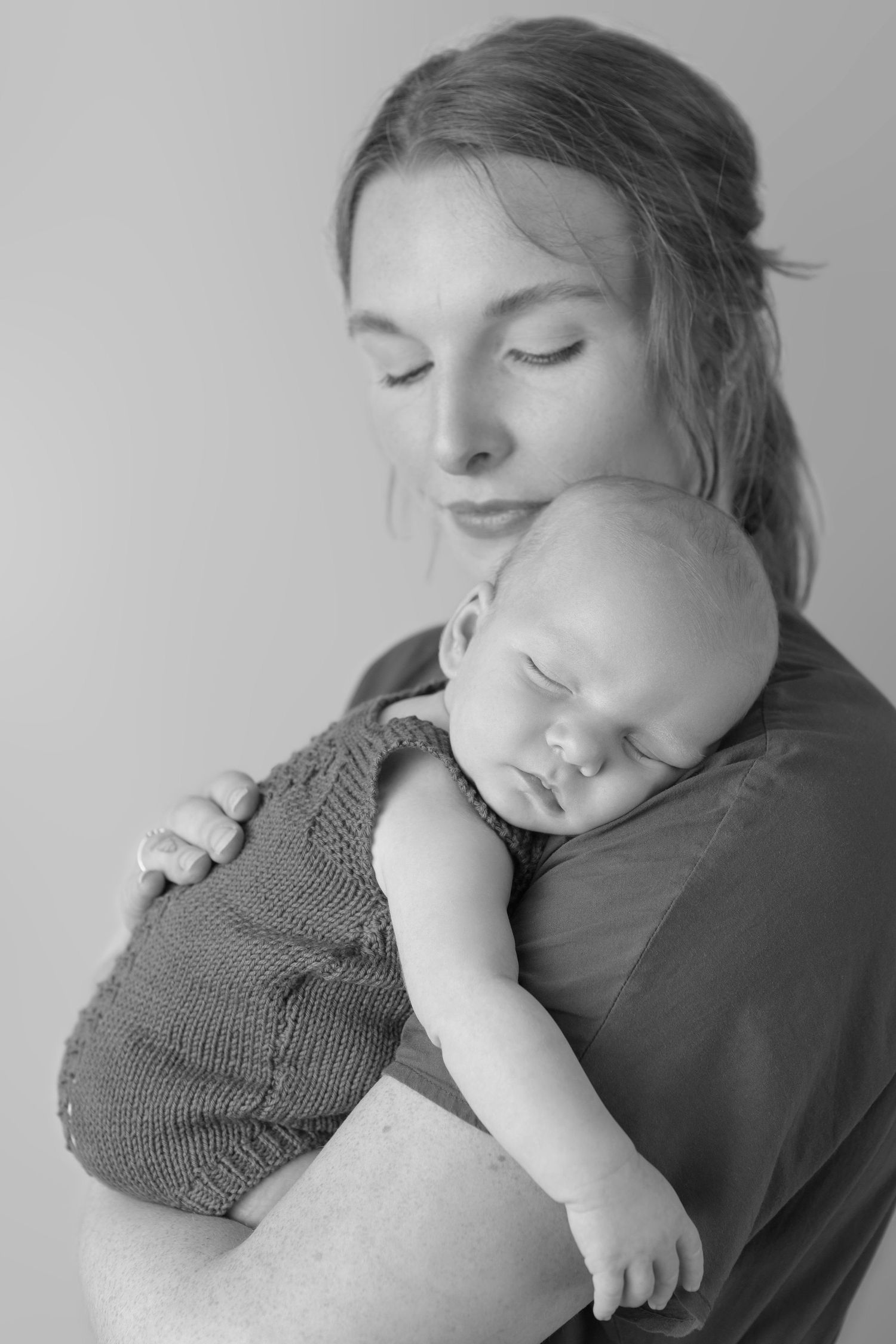 Black and white portrait showing peaceful sleeping baby in knit romper being held against mum's shoulder.