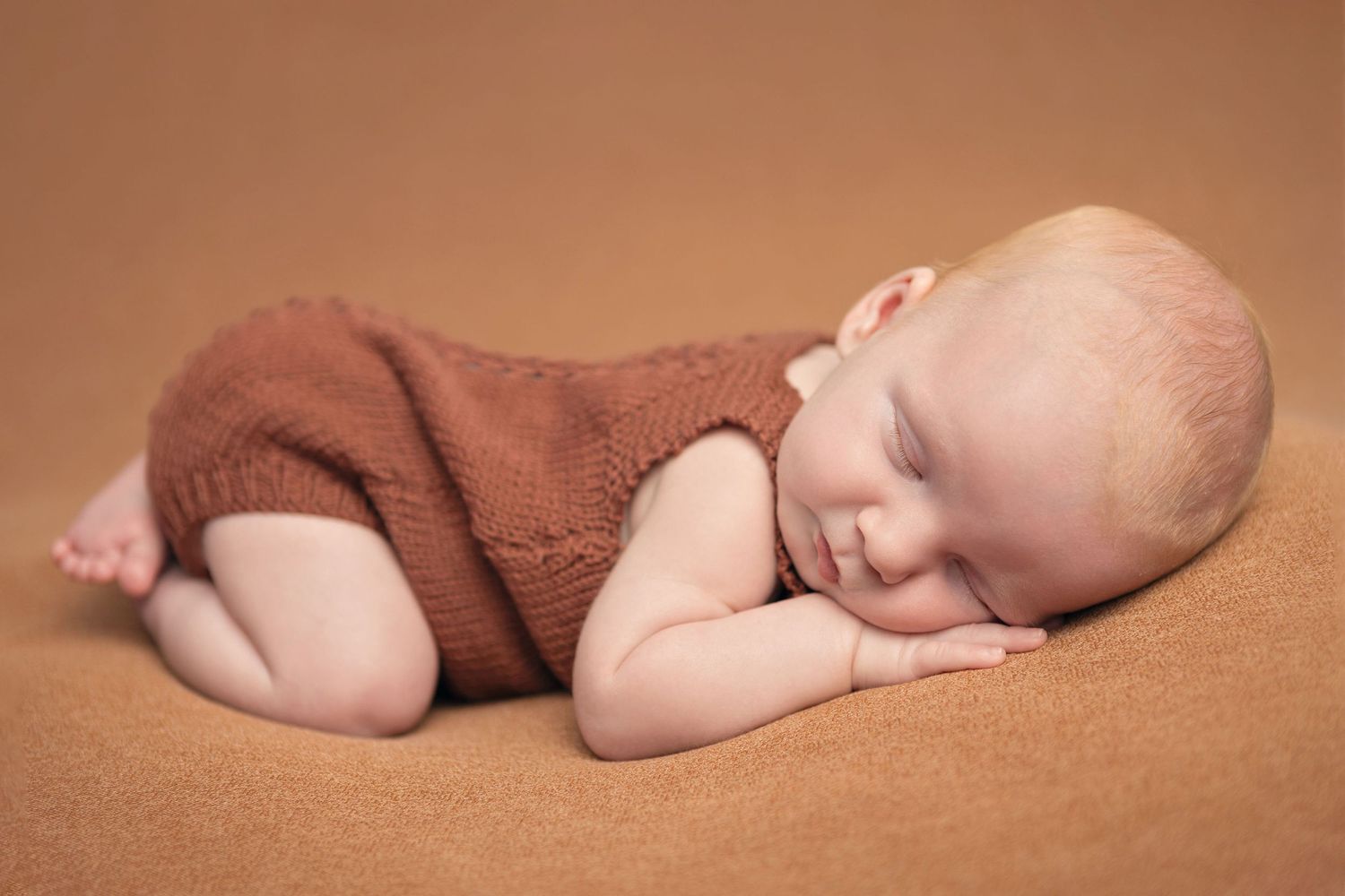 Sleeping newborn wearing brown knit romper lying on textured brown fabric.
