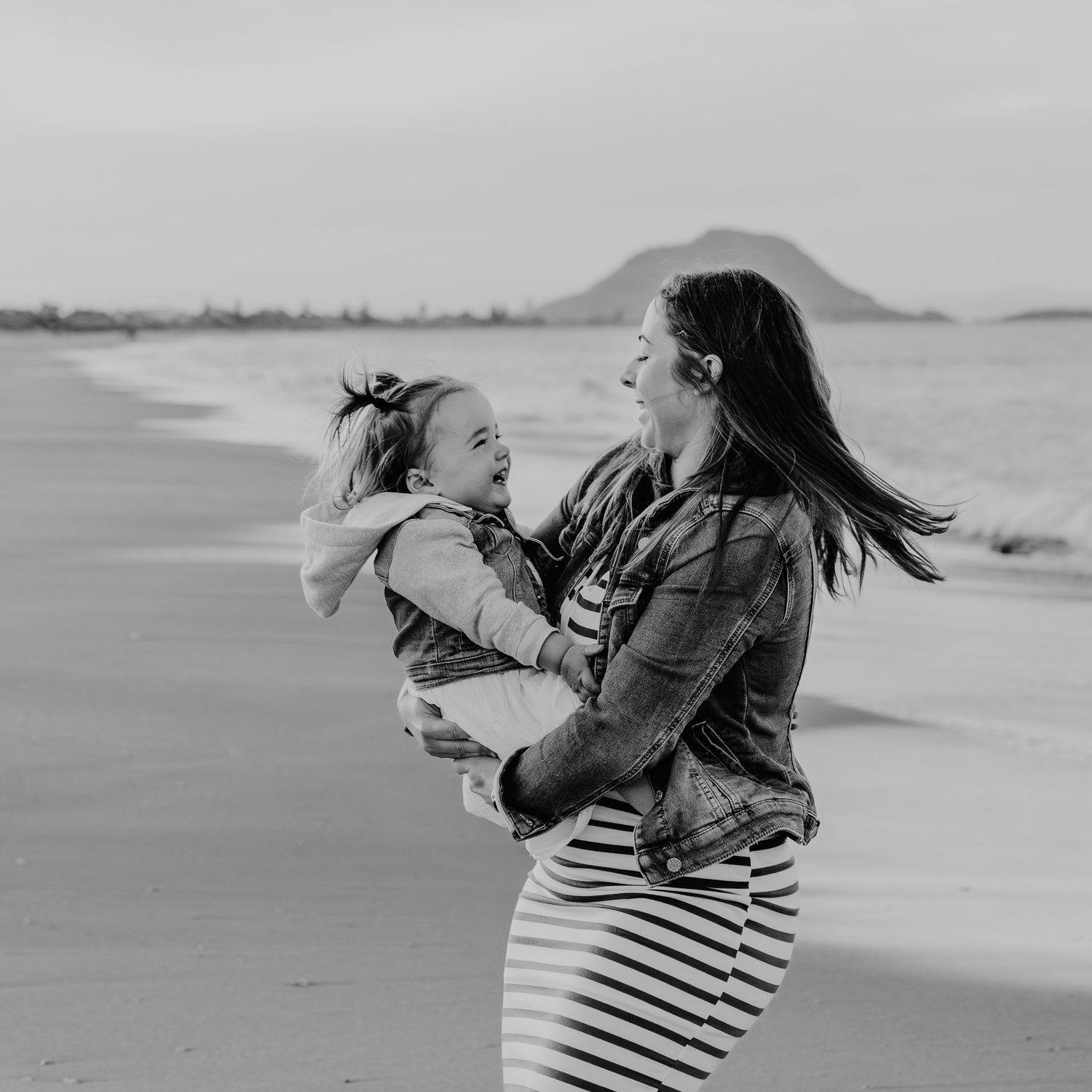 A joyful mother and child share a laugh together on the beach with a mountain visible in the background. Bay of Plenty motherhood photograph