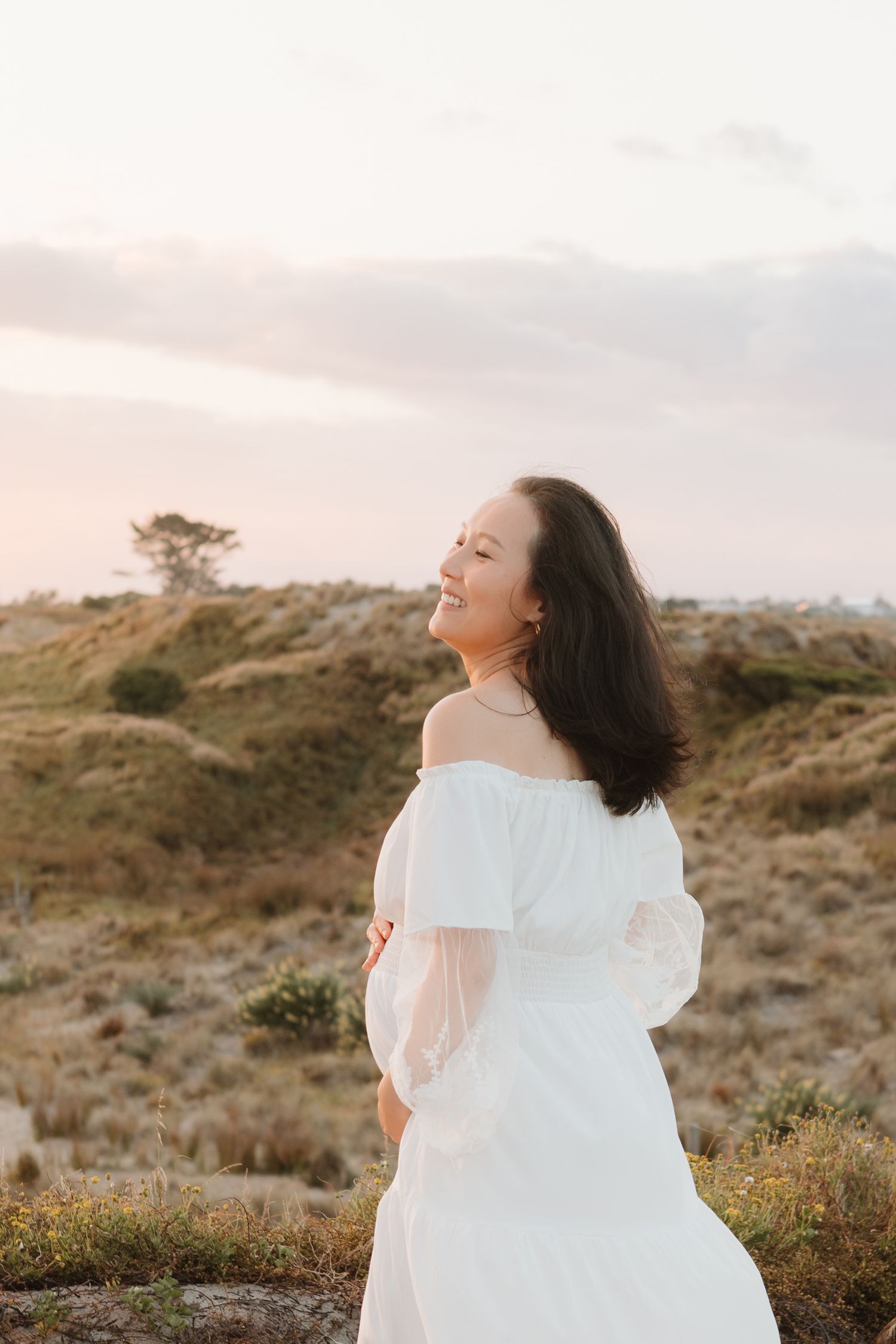 Woman in white dress smiles while standing on a hilltop during sunset.