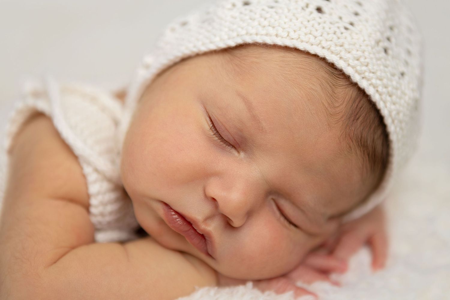 Sleeping baby wearing white knit hat on soft white fabric. Newborn photos Tauranga.