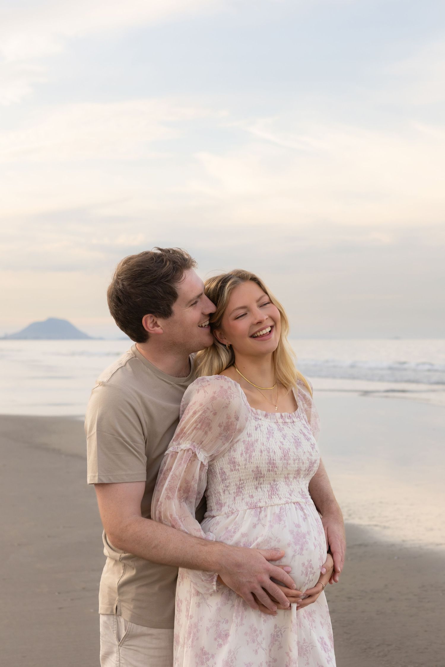 A tender maternity moment captured on a beach at sunset with mountains in the distance.